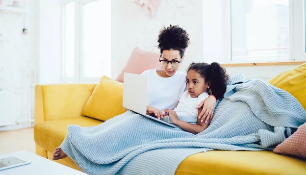 快乐的非洲裔美国女士和惊讶的小女孩坐在舒适的黄色沙发上与笔记本电脑一起在社交媒体上观看视频 — 图库照片