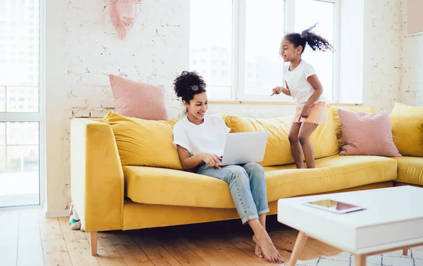 Ethnic daughter jumping near mother in white shirt and jeans sitting on yellow couch and working on laptop at home