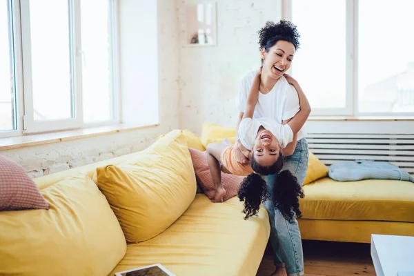 楽しいです正アフリカ系アメリカ人の母親運ぶ幸せな女の子上の手回転周りに持って楽しいですホームで居心地の良いリビングルーム — ストック写真