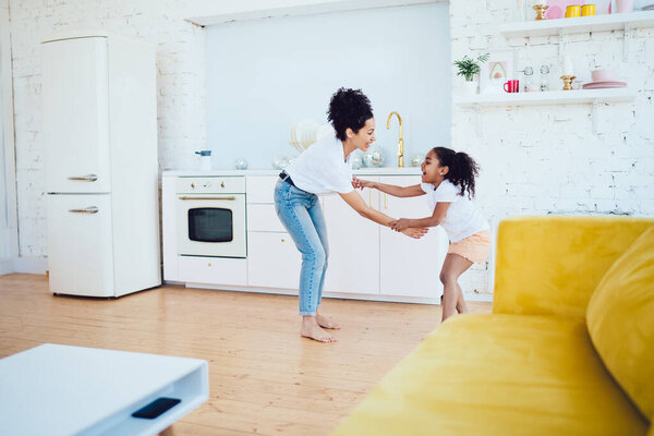 Веселая афроамериканская мать в повседневной одежде с вьющимися волосами, играющая с улыбающейся дочерью, стоя на современной кухне однокомнатной квартиры