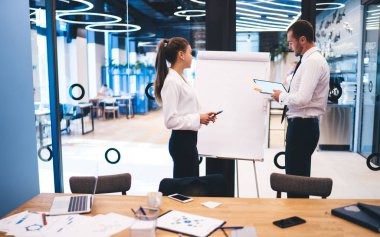 Yaratıcı modern ofiste beyaz tahtanın yanında duran ve sunum sırasında yeni stratejileri tartışan resmi takım elbiseli ciddi meslektaşların yan görünüşü