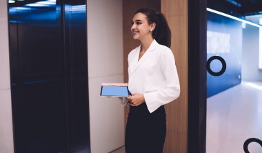 Resmi olarak genç bayan ofis çalışanı elinde dijital tabletle ayakta duruyor ve çağdaş ofis koridorunda gülümsüyor.