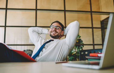 Optik gözlük takan yetenekli girişimci çalışma gününde masa başında modern dizüstü bilgisayarla kameraya gülümsüyor, resmi giyinmiş başarılı ofis çalışanının portresi