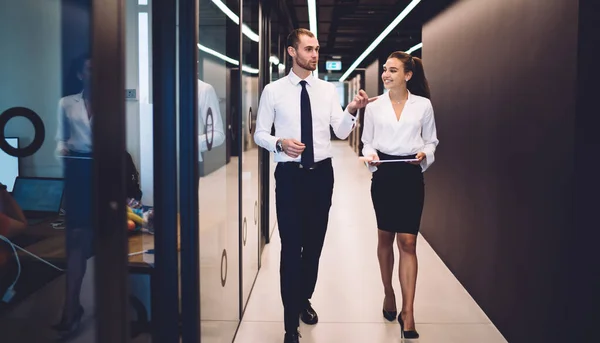 Cheerful young female in heels and formal wear smiling when walking and listening to male colleague while talking funny story at modern workplace