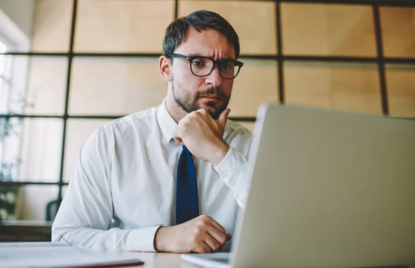 Puzzled bearded entrepreneur checking account balance in online banking service on laptop computer, Caucasian employee in spectacles watching video on website during work time in office interior