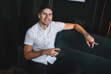 Yukarıdan gülümseyen, cep telefonlu, gerçek kablosuz kulaklıklı, karanlık odadaki kanepede oturan ve hafta sonu dinlenirken kameraya bakan genç bir adamdan.