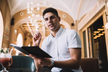 Gündüz vakti modern restoranda oturan defterine notlar yazarken gülümseyen koyu renk saçlı, neşeli bir adam.