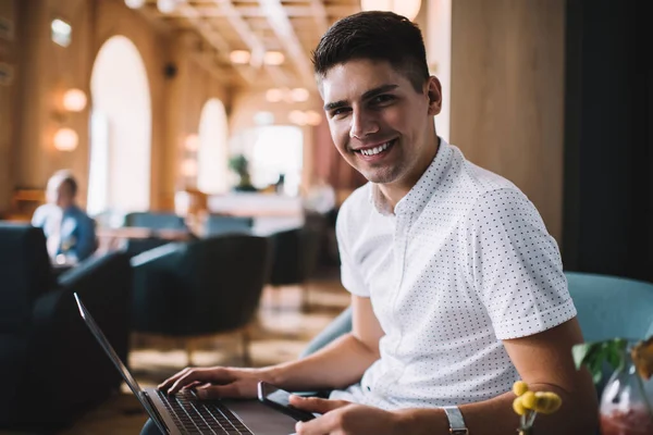 快乐的年轻男性穿着白衬衫 一边浏览笔记本电脑 一边微笑着坐在咖啡桌旁 看着相机 — 图库照片