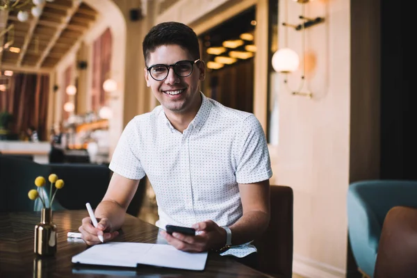 Handsome smiling male student in casual outfit writing essay while using cellphone and making notes at modern restaurant at daytime