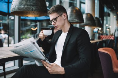 Koyu renkli şık takım elbiseli, gözlüklü, gazete okuyan ve modaya uygun bir kafede elinde bir fincan kahveyle oturan zeki bir erkek.