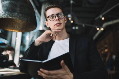 Resmi giyinmiş, gözlüklü, düşünceli bir erkeğin kafede oturmuş gözlerini kaçırırken roman üzerinde çalışması.