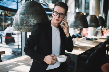 Gözlüklü, iyi giyimli, genç bir erkek. Kafede elinde bir bardak sıcak içecekle dikilirken gözlerini kaçırıp telefonla arıyor.