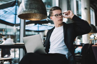 Zarif takım elbiseli ve gözlüklü düşünceli genç adam, masanın yanında dizüstü bilgisayarla oturuyor ve modern kafe projesinde çalışırken gözlerini kaçırıyor.