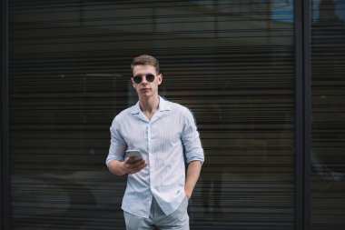 Gündelik giyinen ve güneş gözlüklü genç adam cep telefonuyla internette gezinirken gün ışığında karanlık duvarın arkasında el ele dikiliyor.