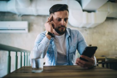Ciddi sakallı bir adam, günlük giysiler içinde, kafeteryada bir bardak suyla, ahşap masada oturuyor ve akıllı telefon kullanıyor.
