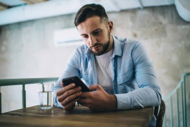 Ciddi sakallı bir adam gündelik giysiler içinde modern kafedeki ahşap masada oturuyor ve akıllı telefon kullanıyor.