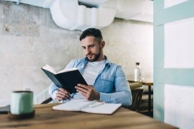 Yüzünde sakallı genç bir adam gündelik giysiler içinde masada otururken kitap okuyor.
