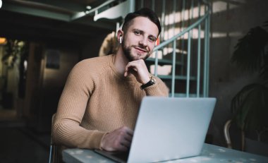 Mutlu karizmatik erkek serbest çalışan, elleri çenesinde rahat bir odada dizüstü bilgisayarla oturuyor ve kameraya bakarken gülümsüyor.