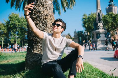 Palmiye ağacının altında yeşil çimlerde oturan ve Barselona 'da yaz tatilinde akıllı telefondan selfie çeken neşeli İspanyol erkek turistin düşük açısı.