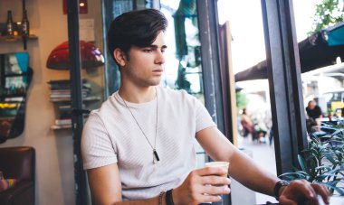 Sakin, beyaz tişörtlü, pencerenin yanında otururken bir bardak kahve içen ve bilgisayarla uzaktan çalışan modern bir erkek.