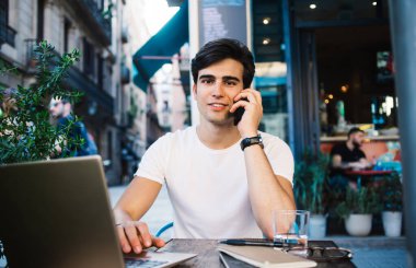 Genç yakışıklı adam, Barcelona 'da bir kafede oturmuş kameraya bakarken dizüstü bilgisayar kullanıyor ve telefonla konuşuyor.