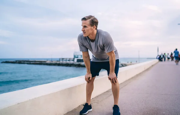 Young caucasian male jogger breathing on pause during running training legs on morning outdoor workout,thoughtful sportsman looking at water standing on quay resting on training keeping healthy body