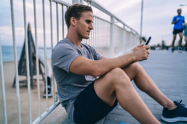 Serious sportsman in active wear sitting outdoors using mobile phone for blogging after training keeping healthy lifestyle, handsome man athlete using app for checking results and reaching goals