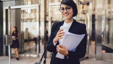 Siyah takım elbiseli, gözlüklü, çantalı ve evraklı neşeli bir kadın New York 'taki modern iş merkezinin girişinin önünde duruyor.