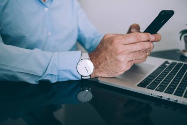 Beyaz kol saati olan, resmi giysili, tanınmayan meşgul bir adamın akıllı telefonuyla etkileşim halindeyken net kitapla yan yana oturduğu yüksek açı.