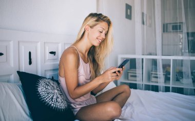 Mutlu kadın blogcu rahat yatak odasında boş zamanlarında çevrimiçi bilgileri kontrol ediyor, cep telefonu uygulaması aracılığıyla ağ mesajlaşması sırasında akıllı telefon iletişiminin keyfini çıkarıyor