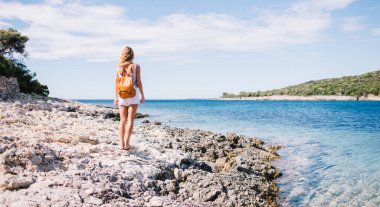 Güneşli bir günde, temiz deniz suyunun yanında sırt çantalı, tanınmayan bir kadının arka görüntüsü.