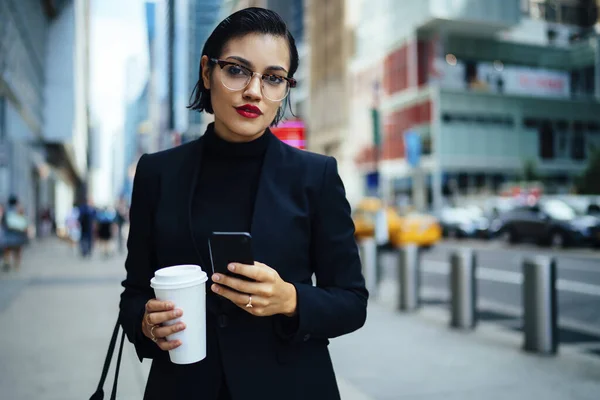 Elegant young woman in formal wear and eyeglasses holding cup of hot drink and looking at camera while standing in modern city centre