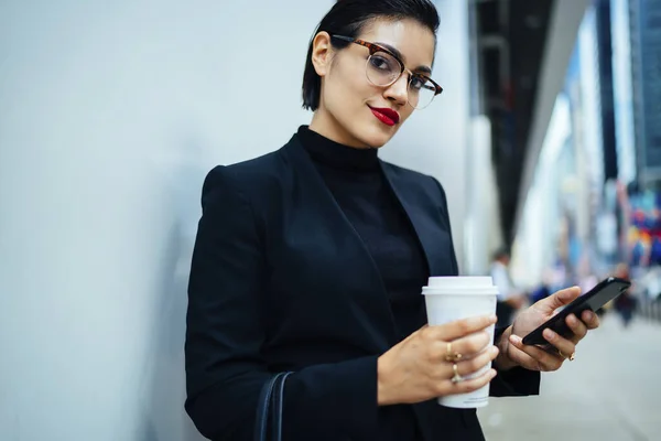 身穿黑色服装 戴着眼镜 留着短发的优雅年轻女性站在墙边 手里拿着纸杯 一边浏览着手机 — 图库照片