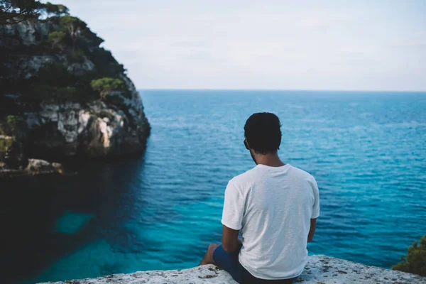 白いTシャツを着た認識できない男性旅行者のバックビューは岩の端に座っており メノルカ島の夏休みの間に海を賞賛 — ストック写真
