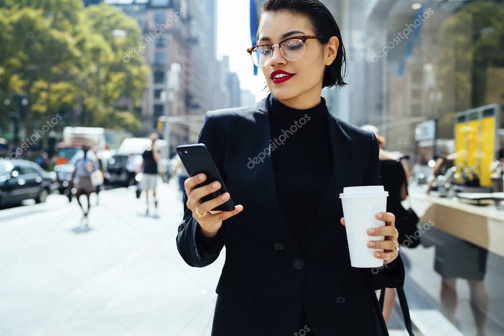 Smiling elegant female in formal wear and eyeglasses with red lipstick messaging on smartphone and drinking takeaway coffee on street of New York City