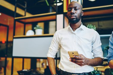 Resmi kıyafet giymiş Afrikalı Amerikalı girişimcinin yarım boy portresi ofis akışı sırasında kameraya bakıyor, esmer tenli iş adamı 30 'lu yaşlarda cep telefonu teknolojisi tutuyor ve şirkette poz veriyor.