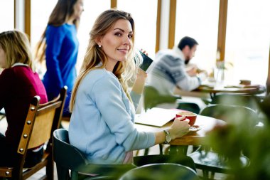 Rahat bir kafede yuvarlak masada oturan ve bir fincan kahve içen gündelik kıyafetli hoş, gülümseyen genç bir kadının yan görüntüsü.
