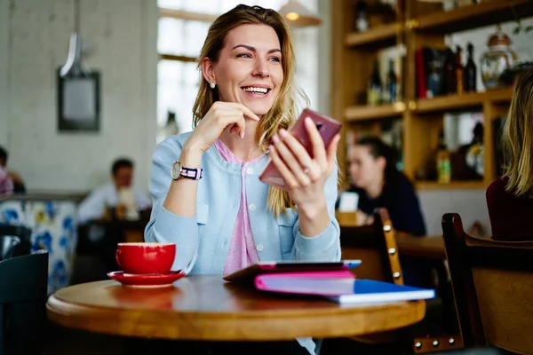 笑容满面 一头金发的年轻女士在咖啡厅里轻松地坐在圆桌边 边喝咖啡边写着便条 并通过智能手机进行交流 — 图库照片