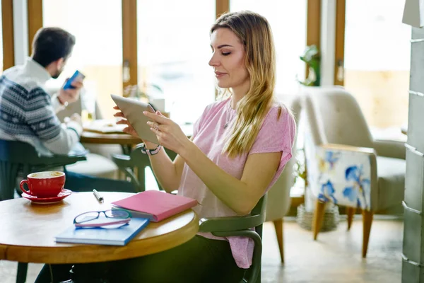 穿着休闲装的积极的年轻女性坐在圆桌边 一边拿着笔记本和咖啡 一边看平板电脑视频 一边在咖啡馆里消磨时光 — 图库照片