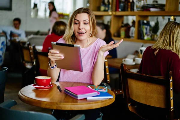 Content  woman in casual clothes smiling while reading results on tablet sitting at wooden table with cup of hot coffee in modern cafe