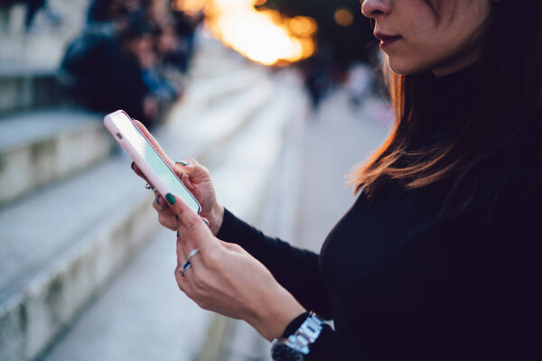Side view of crop female in casual outfit and wristwatch browsing information on smartphone while standing on street near modern buildings