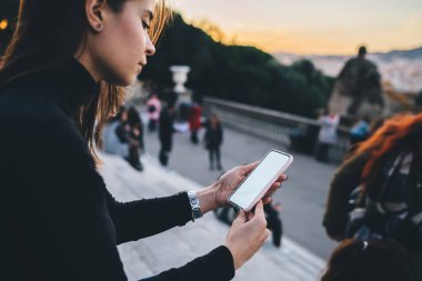 Günbatımında yaya geçidi yakınlarında taş merdivenlerde otururken günlük giysiler içinde esmer, akıllı telefon sörfü yapıyor.