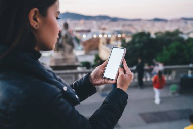 Sıcak ceketli genç bir kadının şehirde vakit geçirmesi ve bulanık şehir manzarasına karşı boş ekran ile akıllı telefona göz atması yan görünüm