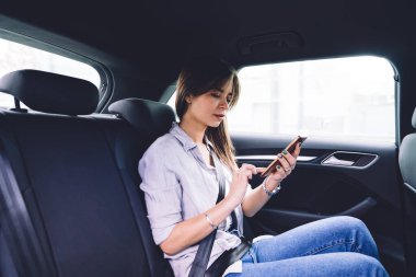 Gündüz vakti arabanın arka koltuğunda emniyet kemerini takmış, akıllı telefonlu, dalgın genç bir kadının yan görüntüsü.