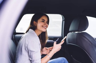 Neşeli gülümseyen, akıllı telefonlu, sıradan elbiseli, yolcu koltuğunda oturan ve mutlu bir şekilde bakışlarını çeviren genç bir kadın.