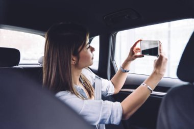 Odaklanmış beyaz tişörtlü genç bir kadının gündüz araba yolculuğu sırasında şehrin akıllı telefonuyla fotoğrafını çekerken yan görüntüsü
