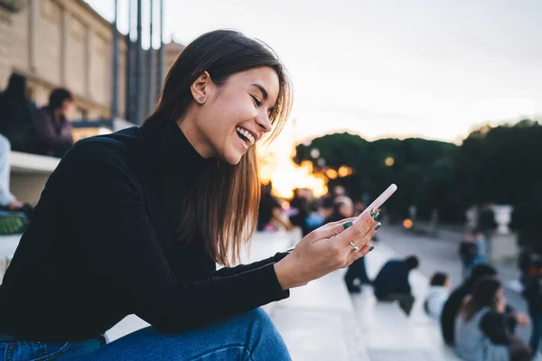 日没時に観光名所の近くの階段で休んでいる間にスマートフォンを使用して笑ってカジュアルな服装で肯定的な若い女性をサイドビュー — ストック写真