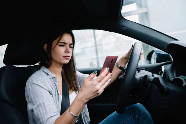 Misunderstand young woman in casual clothes sitting in car and browsing mobile phone while looking at screen on city road