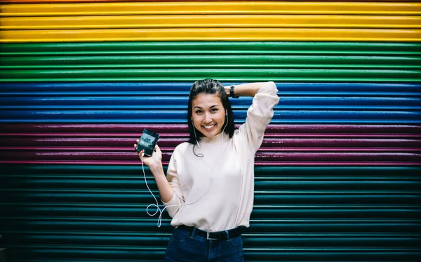 Half length portrait of joyful Asian hipster girl laughing near promotional background using cellphone media app for listening funny audio records, emotional female generation Z in headphones