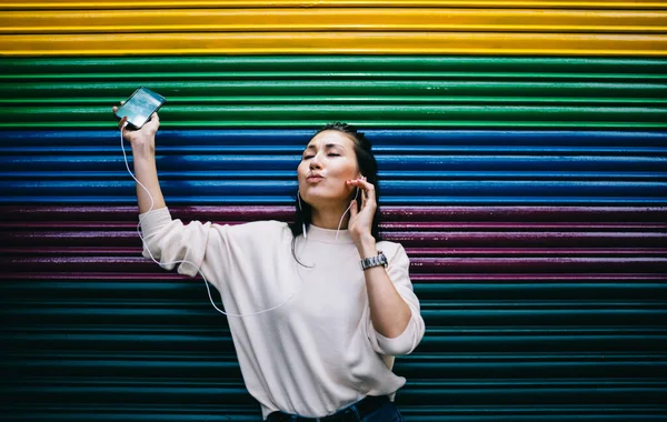 Carefree millennial female in digital headphones enjoying electronic music for dance near publicity wall with copy space area for web media advertising, Asian hipster girl with cellphone having fun